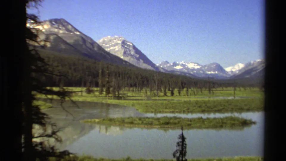 1979 8mm view of a British Columbia mountain valley, with lake water, green shoreline vegetation, trees, and distant snowy peaks.