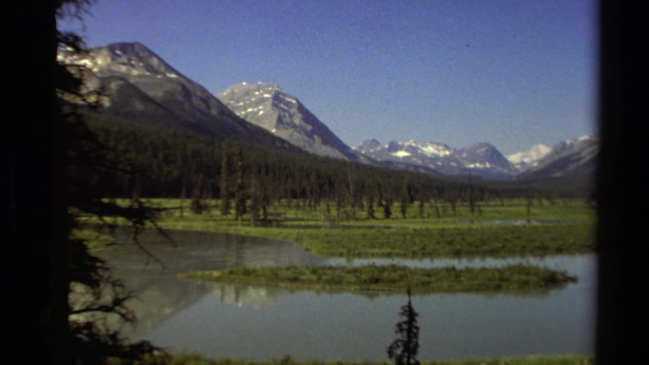1979 8mm view of a British Columbia lake valley with evergreen forest, snow-capped peaks, and dark tree silhouettes at the edges.