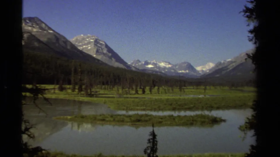 British Columbia mountain valley scene on 8mm film, showing a still lake, green shoreline, evergreen forest, and distant snowy peaks.
