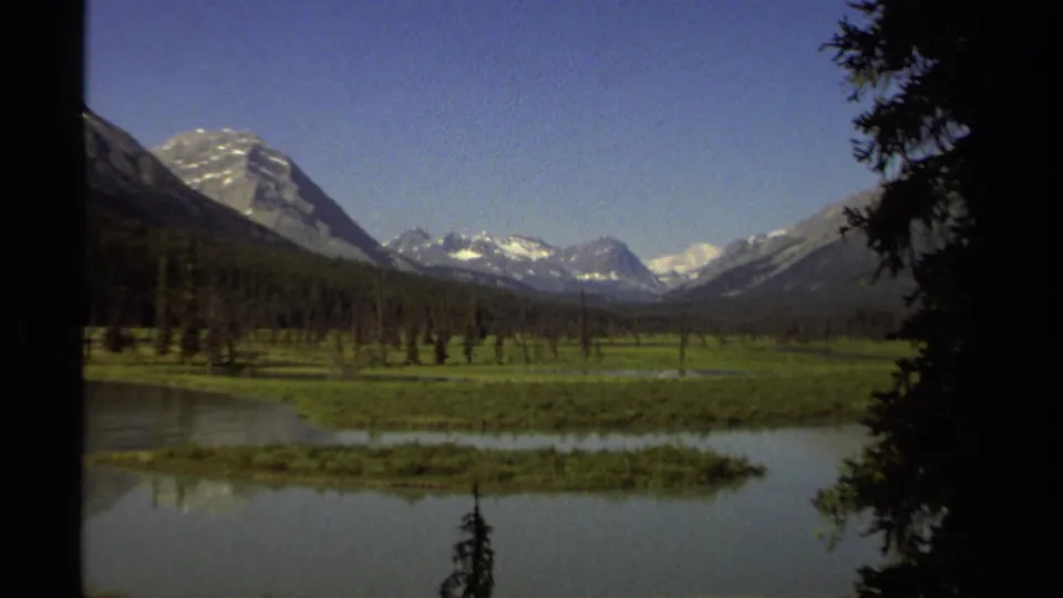 1979 British Columbia landscape frame with dark foreground trees, a reflective lake, green valley floor, and layered snow-capped mountains.
