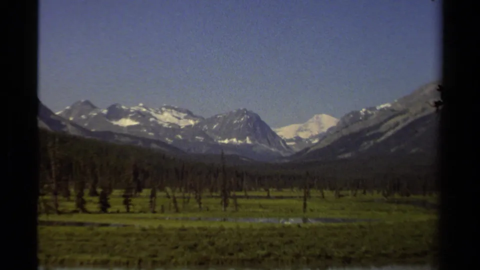 1979 British Columbia landscape showing a broad green valley, thin evergreens, and snow-capped mountain peaks.