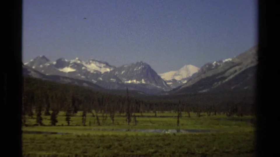 1979 British Columbia 8mm frame of mountains, meadow, scattered trees, and still water in the valley floor.