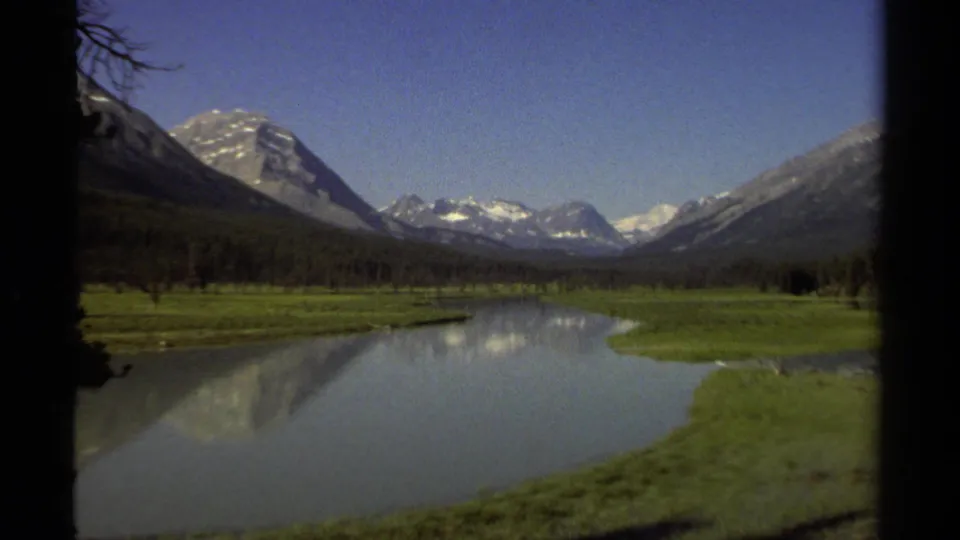 1979 British Columbia view of a calm river reflecting nearby mountains, bordered by green meadow and forest.