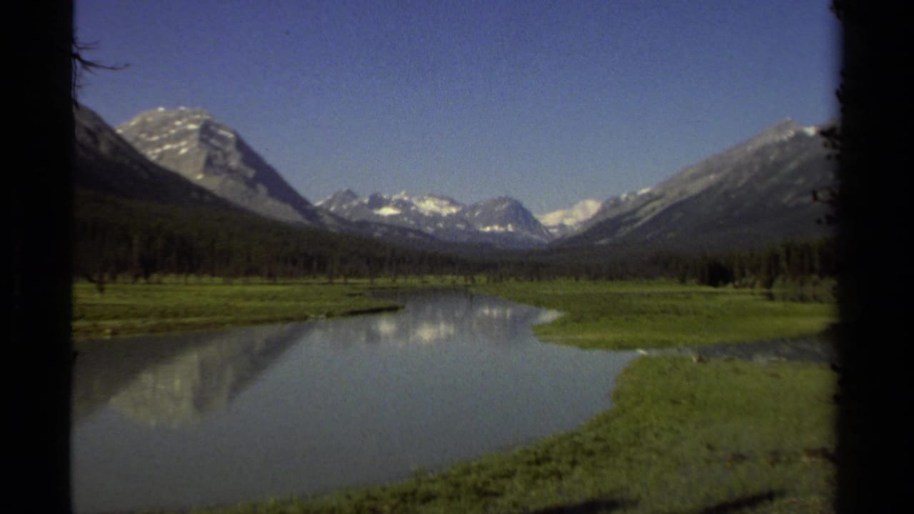 1979 8mm view in British Columbia of snow-capped mountains, blue sky, green riverbanks, and a calm reflective river.