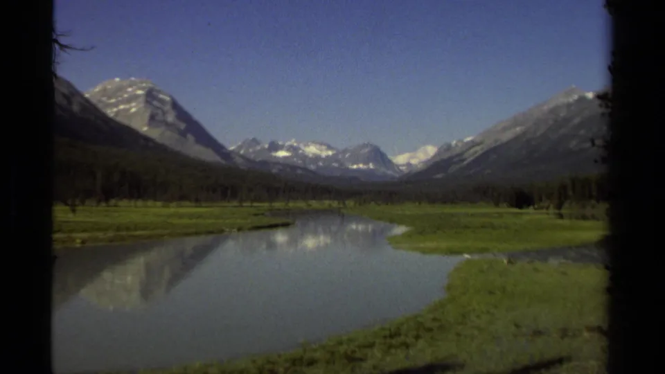 1979 8mm view in British Columbia of snow-capped mountains, blue sky, green riverbanks, and a calm reflective river.