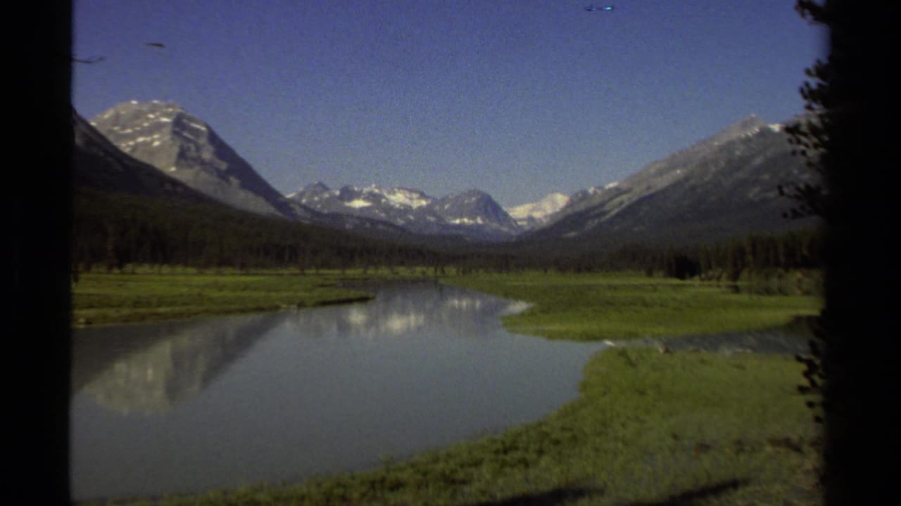 Wide river valley with snow-covered peaks, green banks, and dark trees at the frame edges.