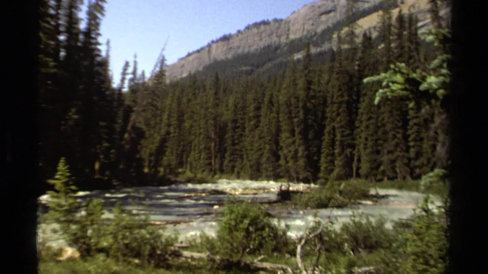 1979 British Columbia home-movie frame showing a sunlit rocky stream below evergreen forest and a distant mountain slope.