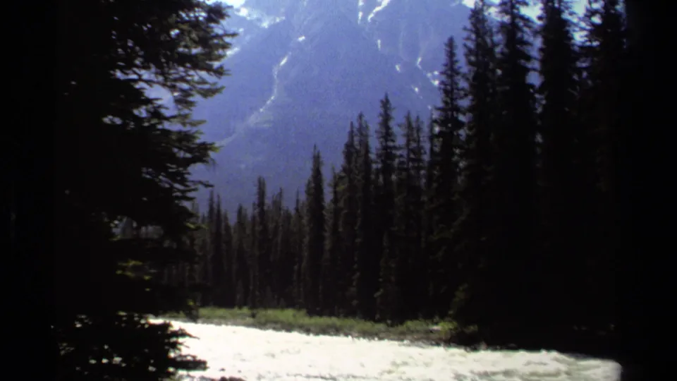 1979 British Columbia 8mm frame with a white river channel, dense conifers, and hazy snow-marked mountains in back.