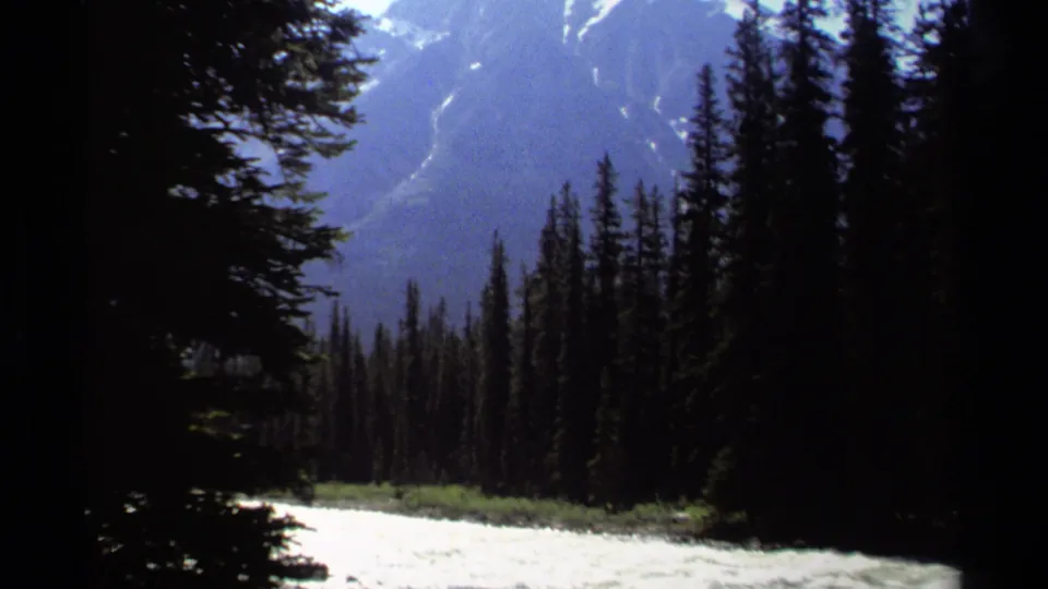 1979 British Columbia 8mm frame showing evergreen trees beside a bright mountain river and blue-toned slopes beyond.