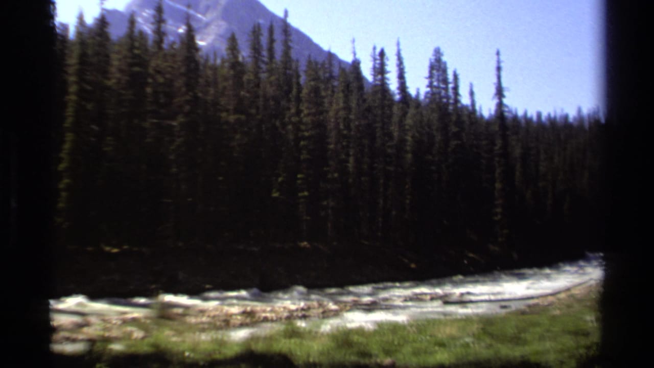 1979 British Columbia 8mm frame showing whitewater river, evergreen trees, grassy bank, and a distant mountain slope.