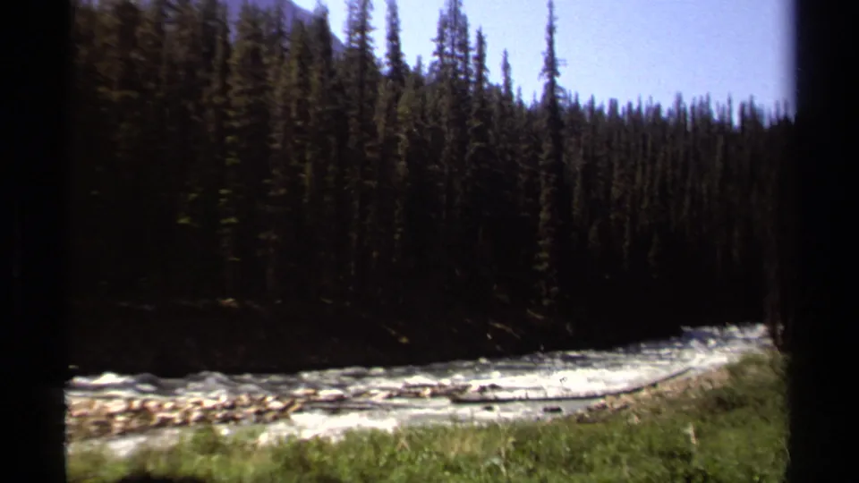 1979 British Columbia home-movie frame of a fast white river running beside conifers and a rocky shoreline.