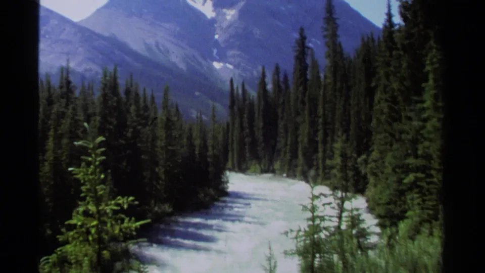 1979 8mm view of a British Columbia river valley framed by spruce-like evergreens and distant blue mountains.
