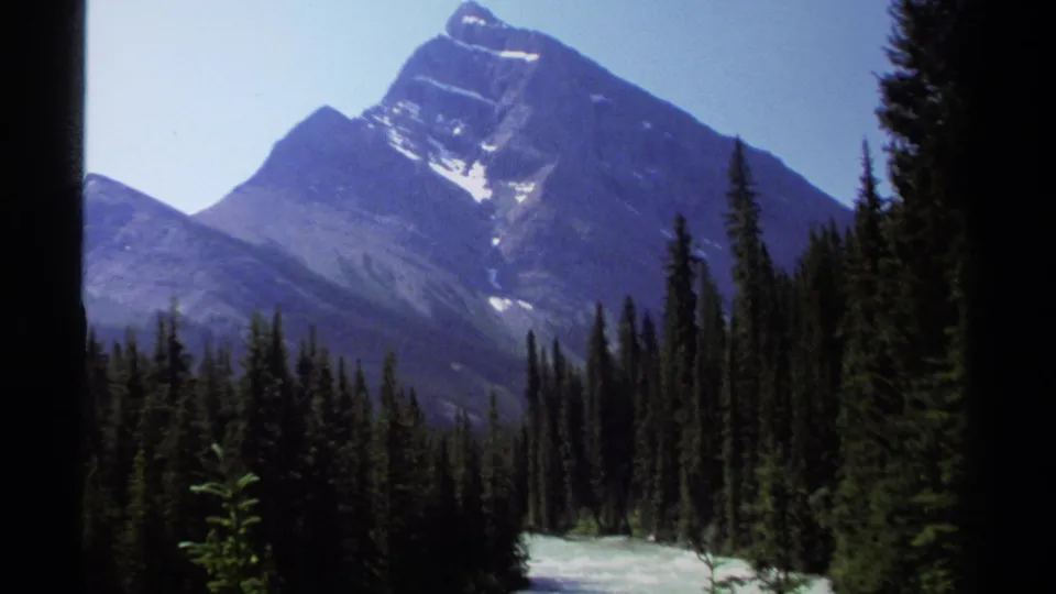 1979 British Columbia home-movie frame showing a snow-streaked mountain peak above forest and river valley.