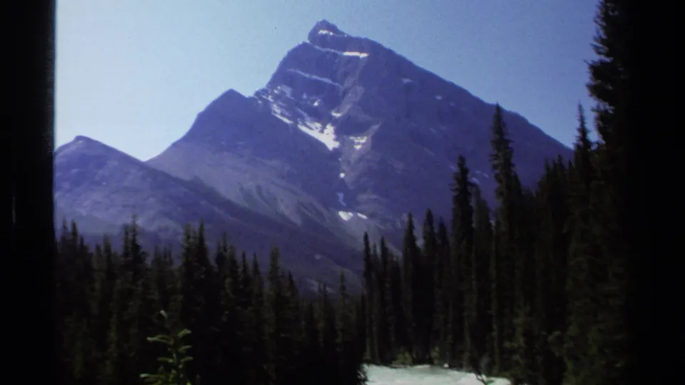 1979 British Columbia 8mm frame of a snow-streaked mountain peak above dense conifer forest under a pale blue sky.