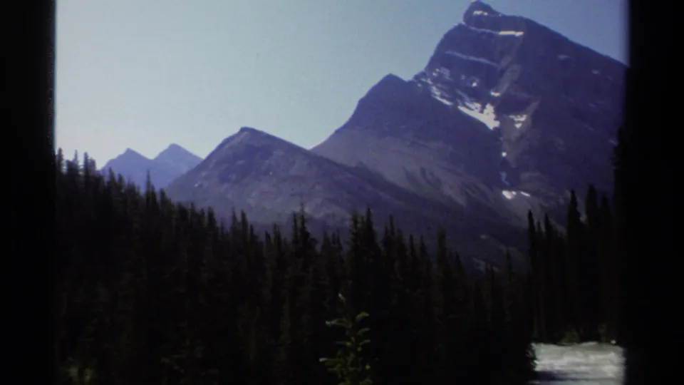 1979 British Columbia 8mm frame showing mountain ridges beyond dense evergreens, with a small area of rushing water at lower right.
