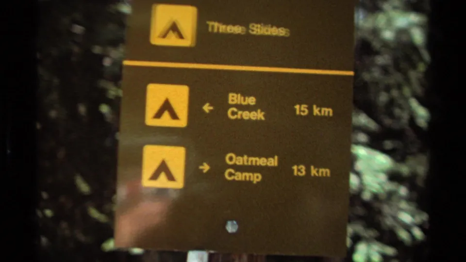 Close view of a forest trail sign in British Columbia, with yellow markers and distances to Blue Creek and Oatmeal Camp.