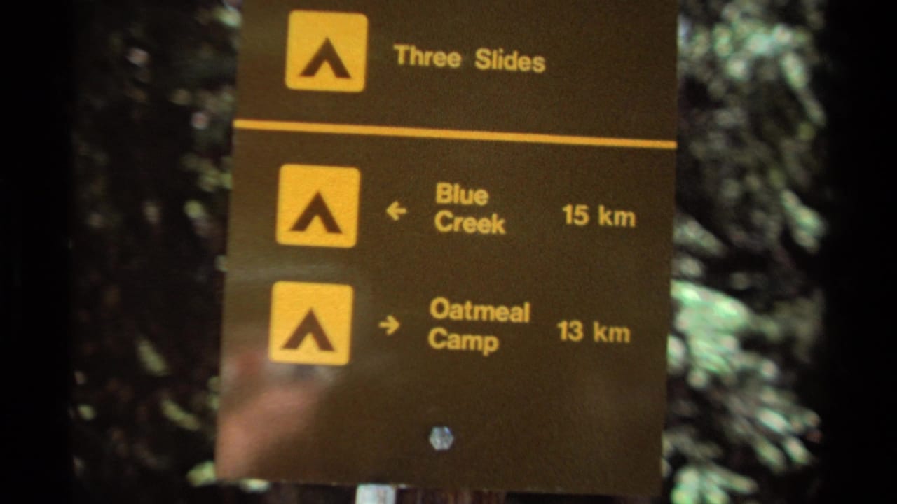 Brown trail marker listing Three Slides, Blue Creek, and Oatmeal Camp in a forest.