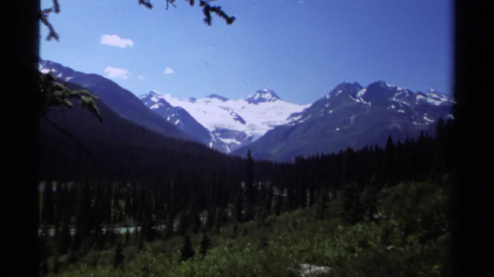 1979 British Columbia 8mm frame showing a wide alpine valley, dark evergreen slopes, snow-capped mountain peaks, and clear blue sky.