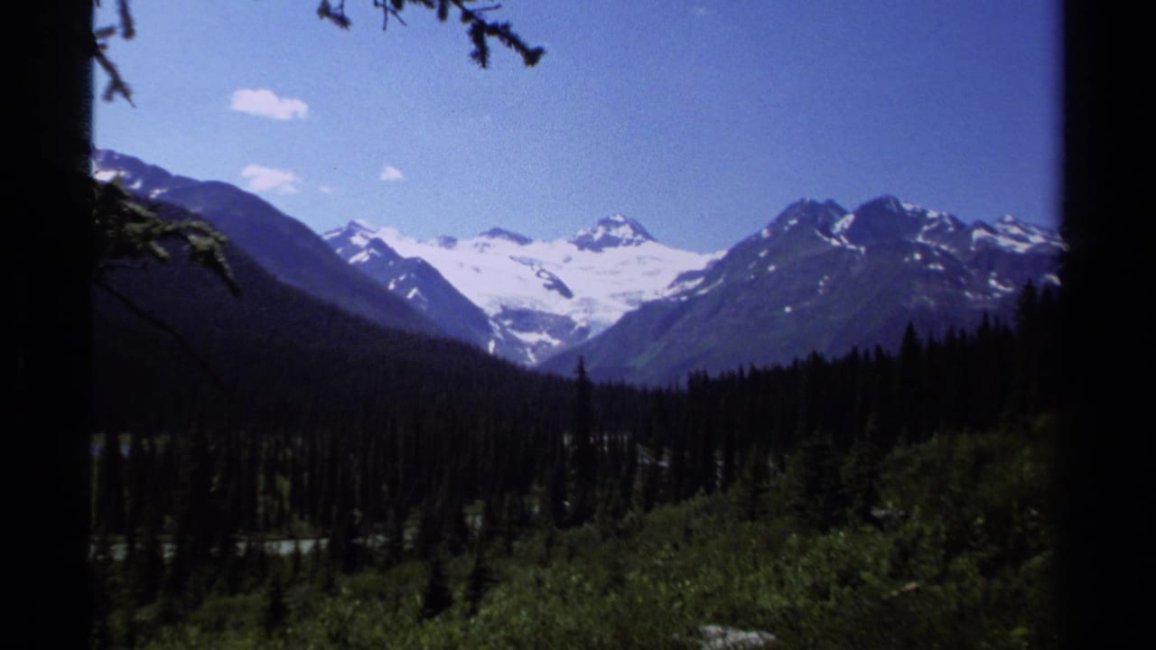 1979 8mm view of British Columbia snow-capped mountains and glaciers beyond a dark evergreen valley.