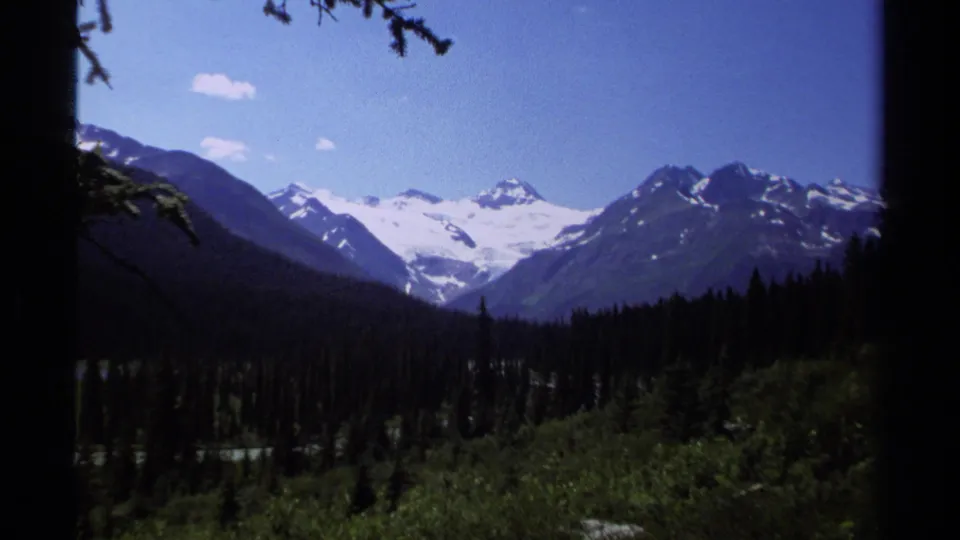 1979 8mm view of British Columbia snow-capped mountains and glaciers beyond a dark evergreen valley.