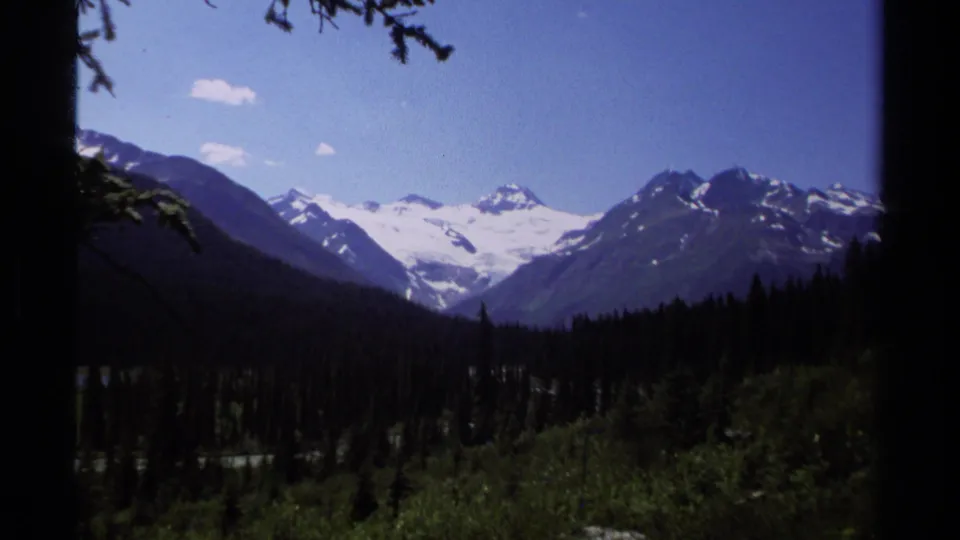 British Columbia alpine landscape on 8mm film, with glacier-covered peaks above dense evergreen forest.