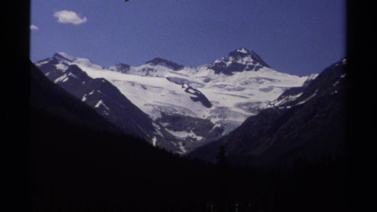 1979 8mm view of snow-capped mountain peaks in British Columbia, with dark forested slopes and a small cloud in blue sky.