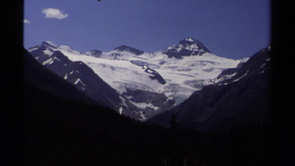 1979 8mm view of snow-capped mountain peaks in British Columbia, with dark forested slopes and a small cloud in blue sky.