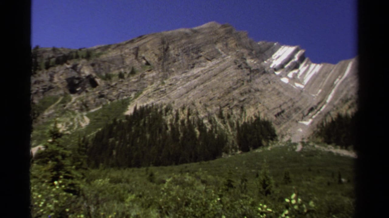 Layered gray mountain ridge with snow patches above green valley and evergreen trees.