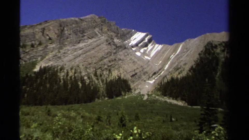 1979 8mm frame of a British Columbia alpine slope, showing gray layered rock, snow streaks, evergreens, and green valley vegetation.