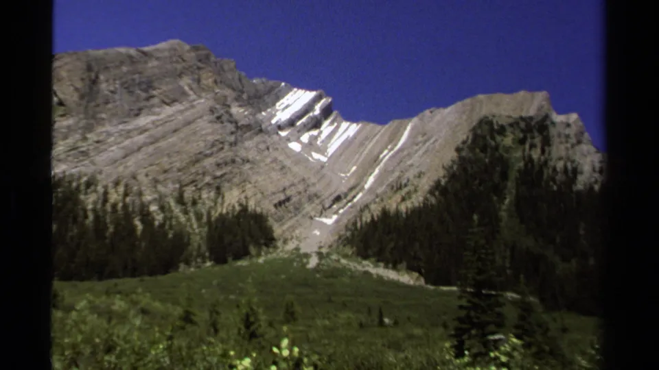 British Columbia alpine landscape from 1979 8mm footage, with gray ridgelines, snow patches, dark evergreens, and green valley floor.