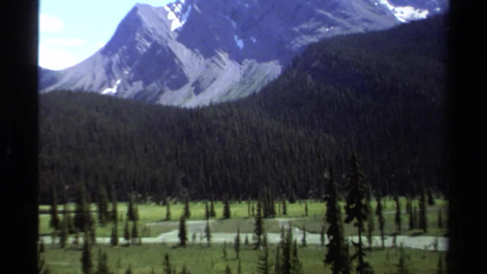 1979 British Columbia home-movie frame showing evergreen forest, meadow-like valley floor, winding water, and distant rocky peaks.