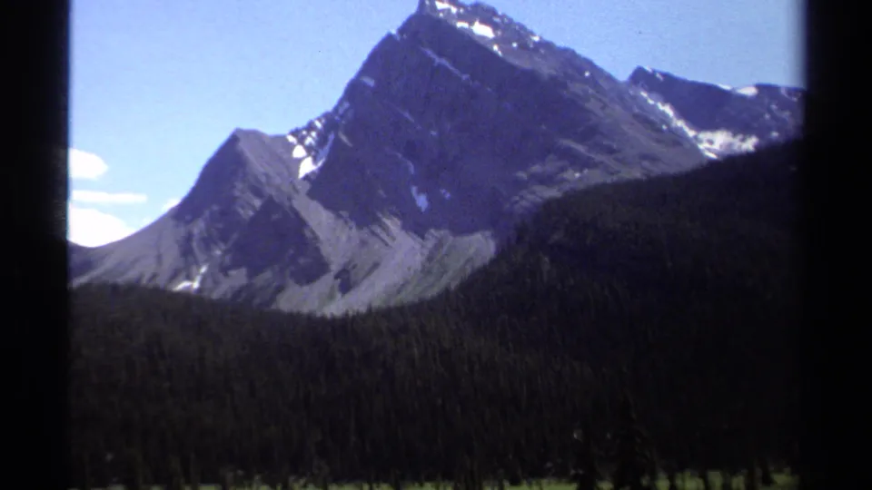 1979 British Columbia 8mm frame with a rocky mountain face rising above dark evergreen forest and a small strip of valley below.