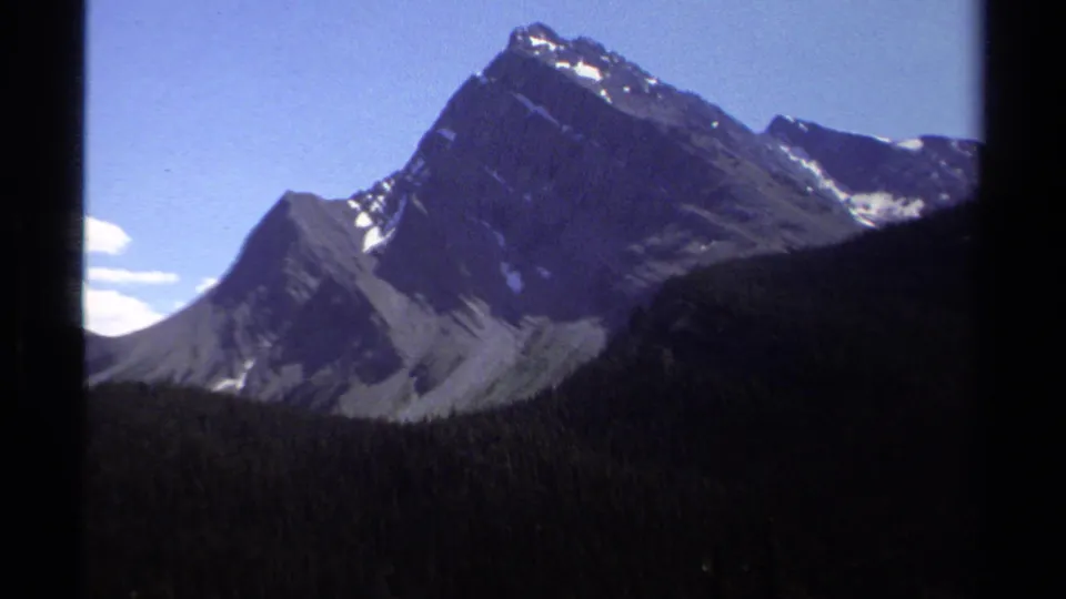 1979 British Columbia home-movie frame focusing on a rugged mountain peak above a dark forested slope, with visible 8mm softness.