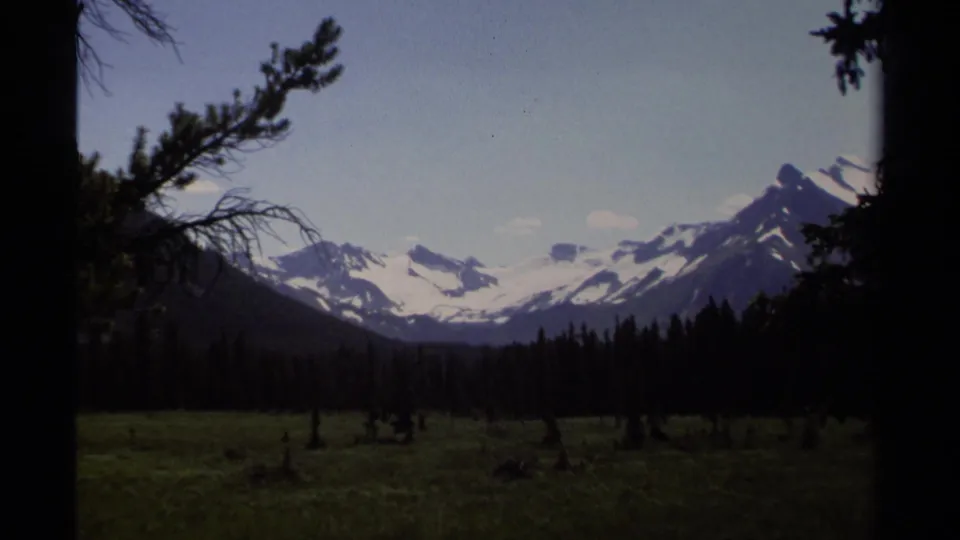 1979 British Columbia 8mm view of snowy mountain ridges across a dark tree line and open meadow.