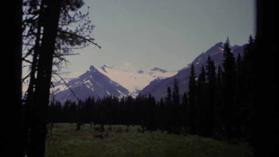 1979 British Columbia alpine view with a pointed snowy peak, dark forest silhouettes, and meadow foreground.