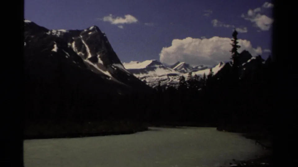 1979 British Columbia river valley view with forested banks, rugged mountains, snowfields, and a large cloud over the peaks.