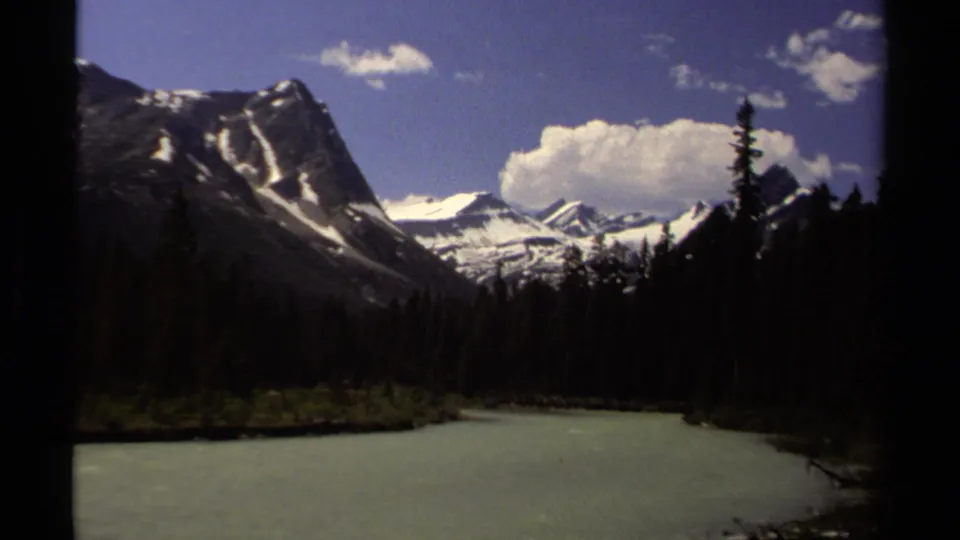 8mm British Columbia landscape with a winding river, conifer banks, dark mountain faces, snowfields, and blue sky.