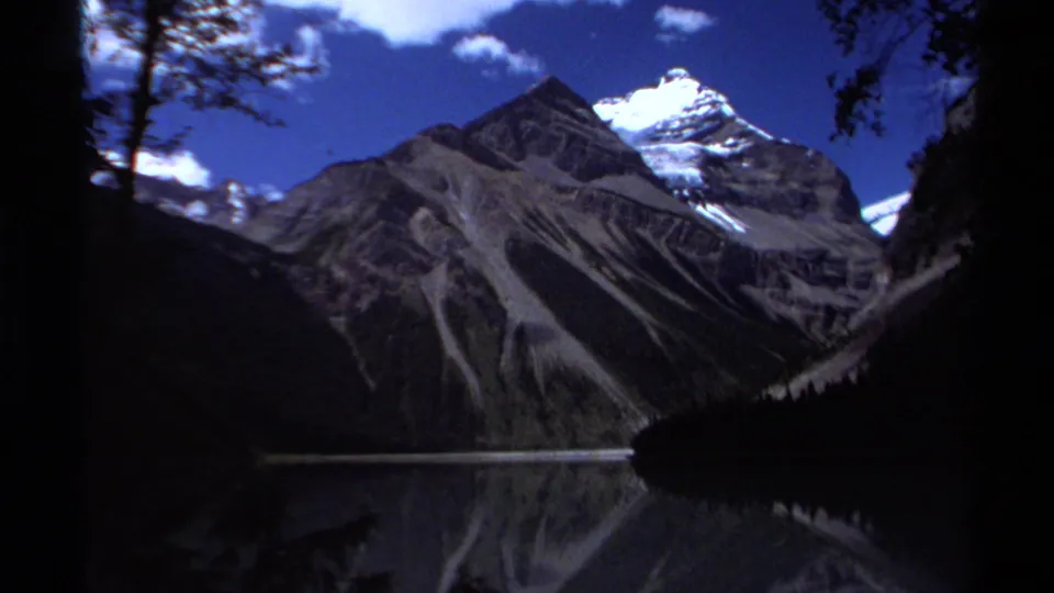 A British Columbia mountain lake appears with steep peaks, snow patches, blue sky, and dark shoreline reflections in 1979 8mm travel footage.