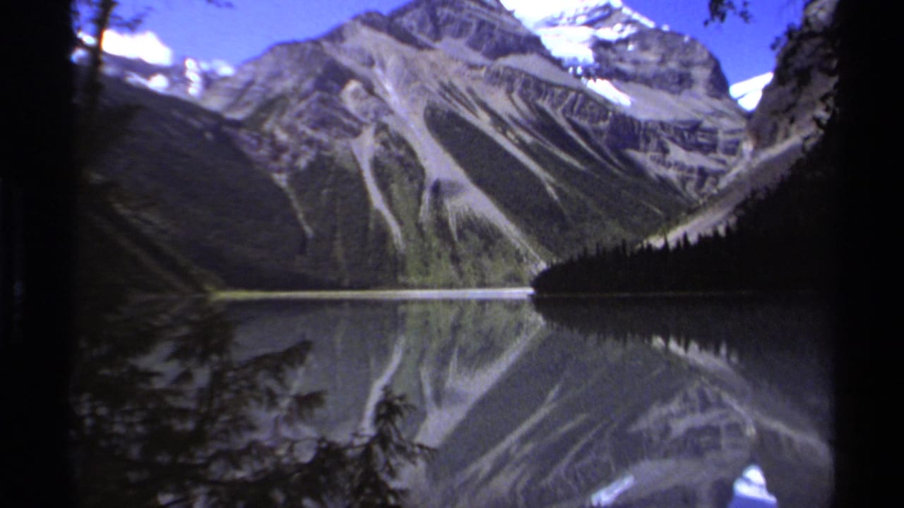 1979 8mm home-movie frame from British Columbia showing rugged mountain slopes and snow patches reflected in a calm lake, with dark evergreen silhouettes at the edges.