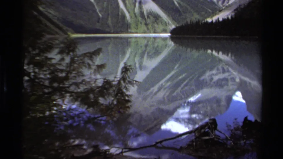1979 British Columbia 8mm frame emphasizing the lake reflection, with dark branches and shoreline vegetation crossing the foreground beneath the mountain slopes.