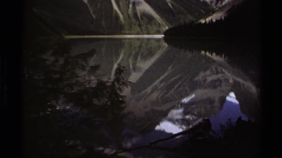 Dimmer 8mm frame from the British Columbia mountain lake sequence, showing reflected slopes, snow patches, and silhouetted trees around the waterline.