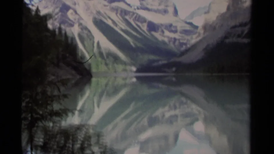 British Columbia 1979 8mm mountain-lake view with rocky peaks, snow patches, conifer shoreline, and soft reflections in still water.