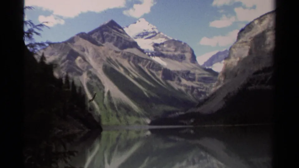 Wide 8mm view of British Columbia mountain scenery in 1979, showing steep rocky slopes, snow patches, still water, and pale clouds.