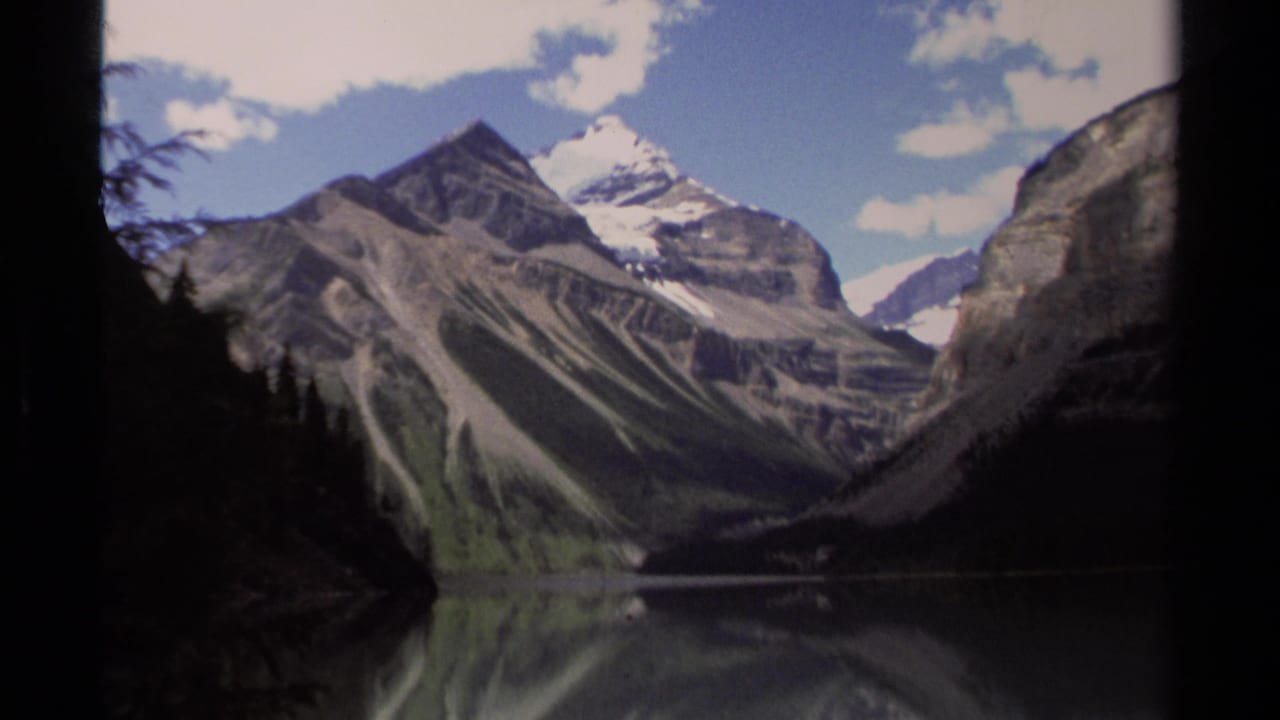 Snow-capped mountains and rocky slopes reflected in a calm lake, framed by dark evergreen trees.