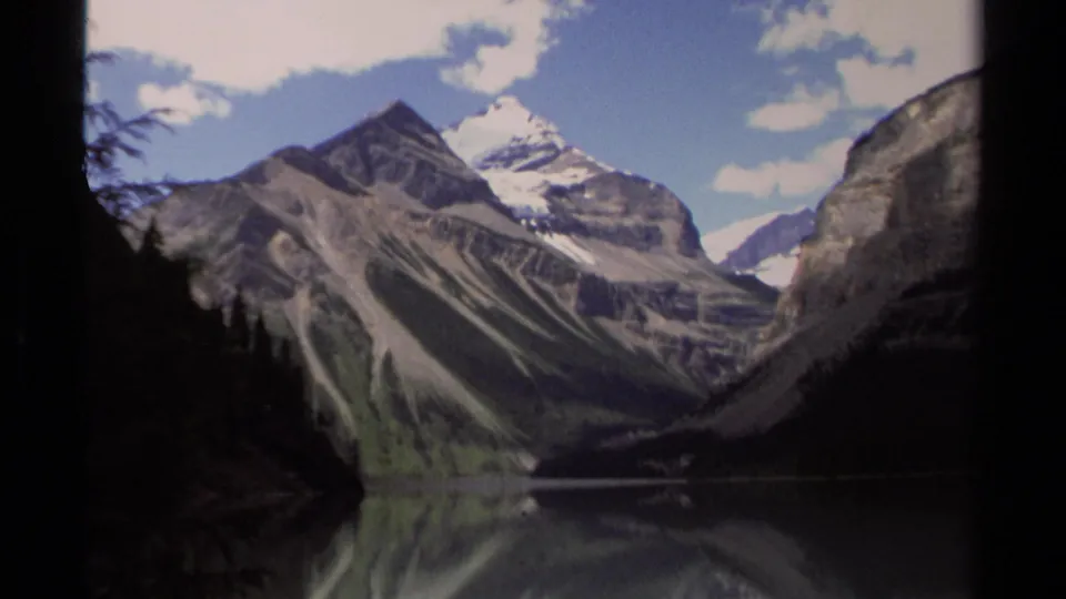 1979 British Columbia 8mm frame of snow-topped mountains and steep slopes above a still lake, with evergreens darkening the foreground edges.