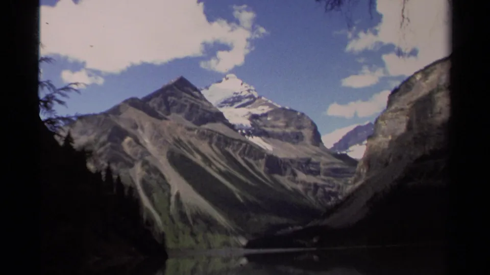 1979 British Columbia scenic frame showing layered mountains, snow patches, a quiet lake, and dark evergreen silhouettes at the sides.