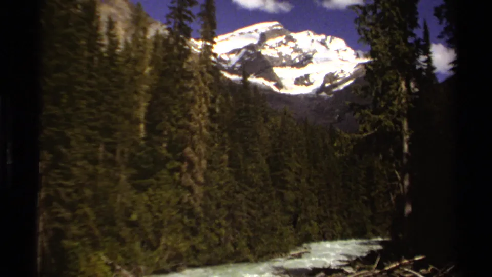 1979 British Columbia mountain landscape with tall evergreens, snow on the peak, and river water low in frame.