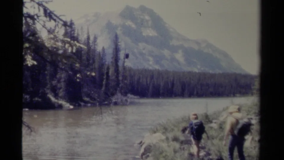 1979 British Columbia home-movie frame showing two hikers near a lake or river, with shoreline brush, evergreens, and snowy mountains behind.