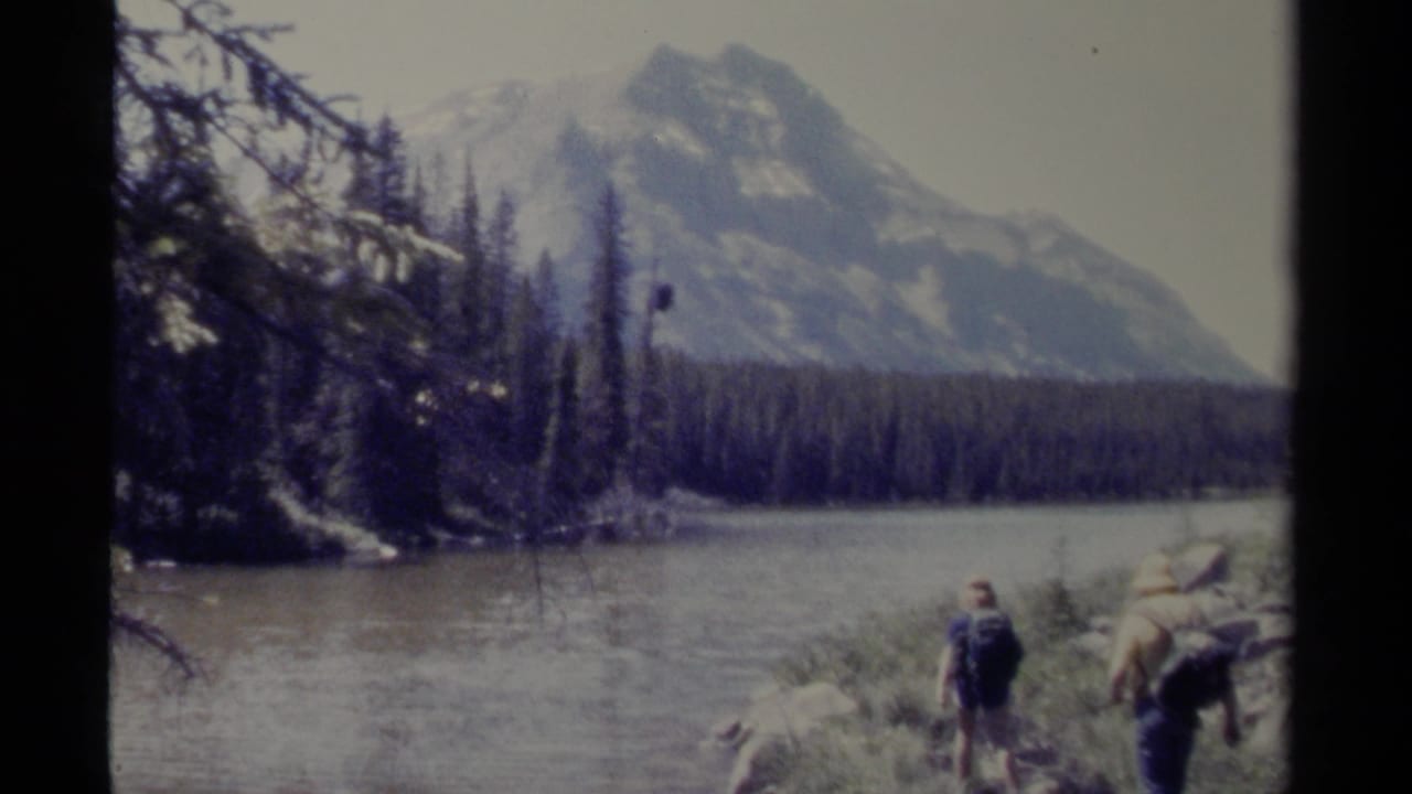 1979 British Columbia 8mm frame showing two hikers by a mountain river or lake, with forest and snow-marked peaks in the background.
