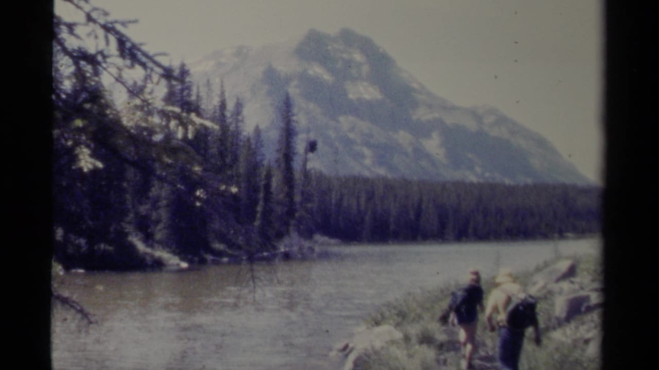 Two distant hikers with packs walk along a British Columbia mountain shoreline in a soft, grainy 8mm home-movie frame.